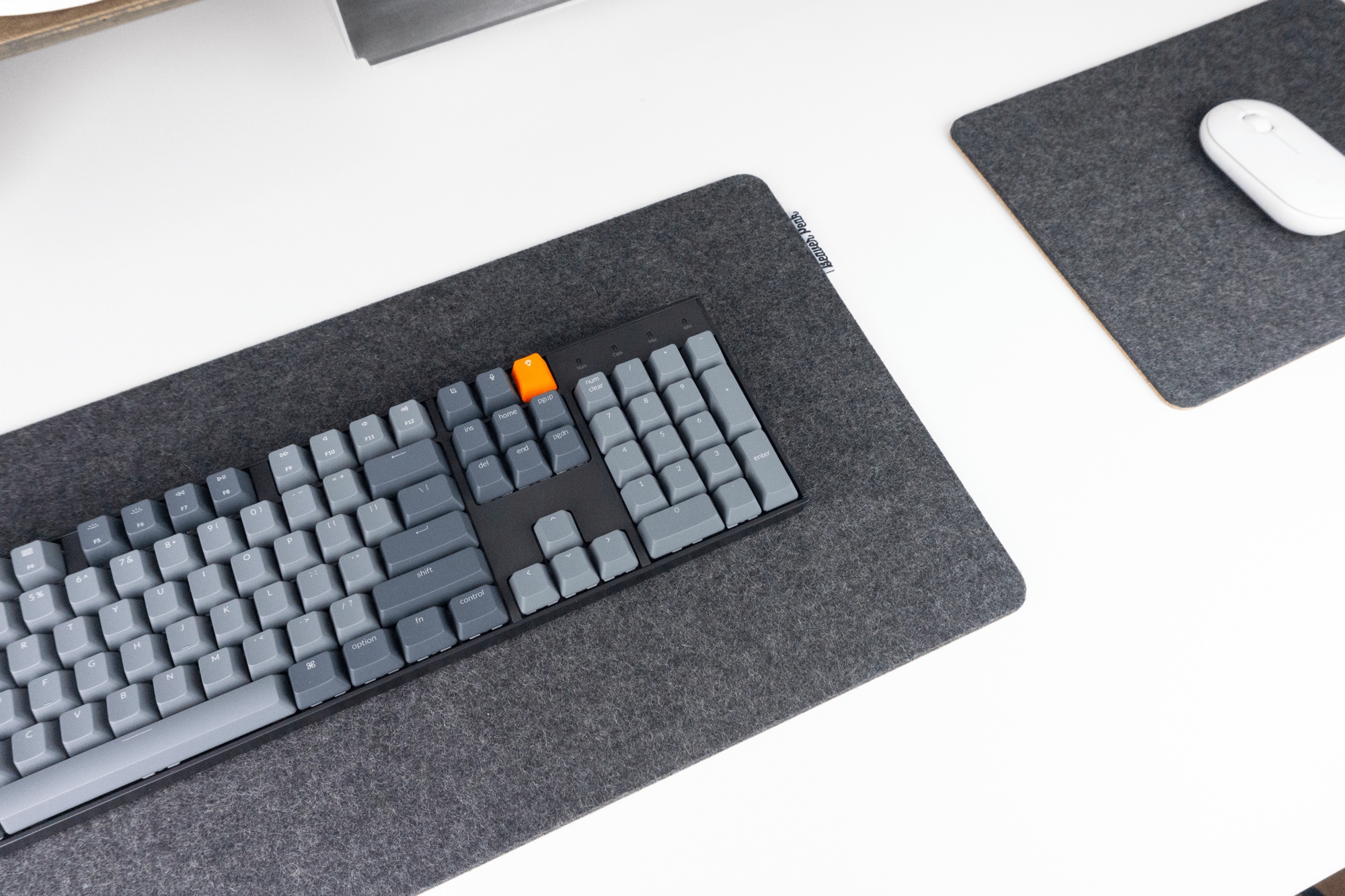 Black BeaverPeak wool and cork desk mat with keyboard on top next to wool and cork mouse pad with white mouse on top. All resting on a white desk top with a monitor stand in the background.