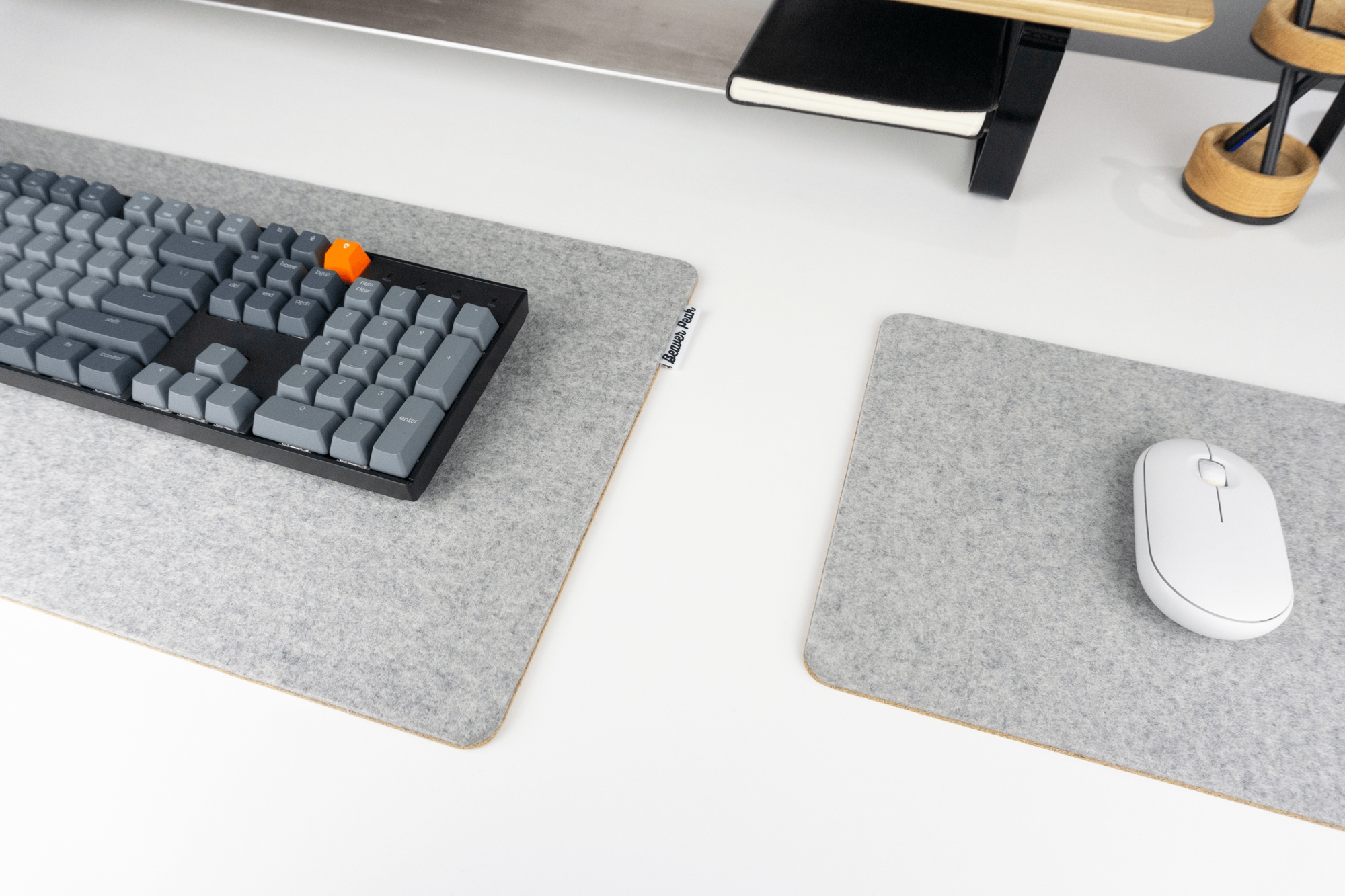 Grey BeaverPeak wool and cork desk pad with keyboard on top next to grey wool felt and cork mouse pad with white mouse on top. All resting on a white standing desk with an oak desk shelf in the background.