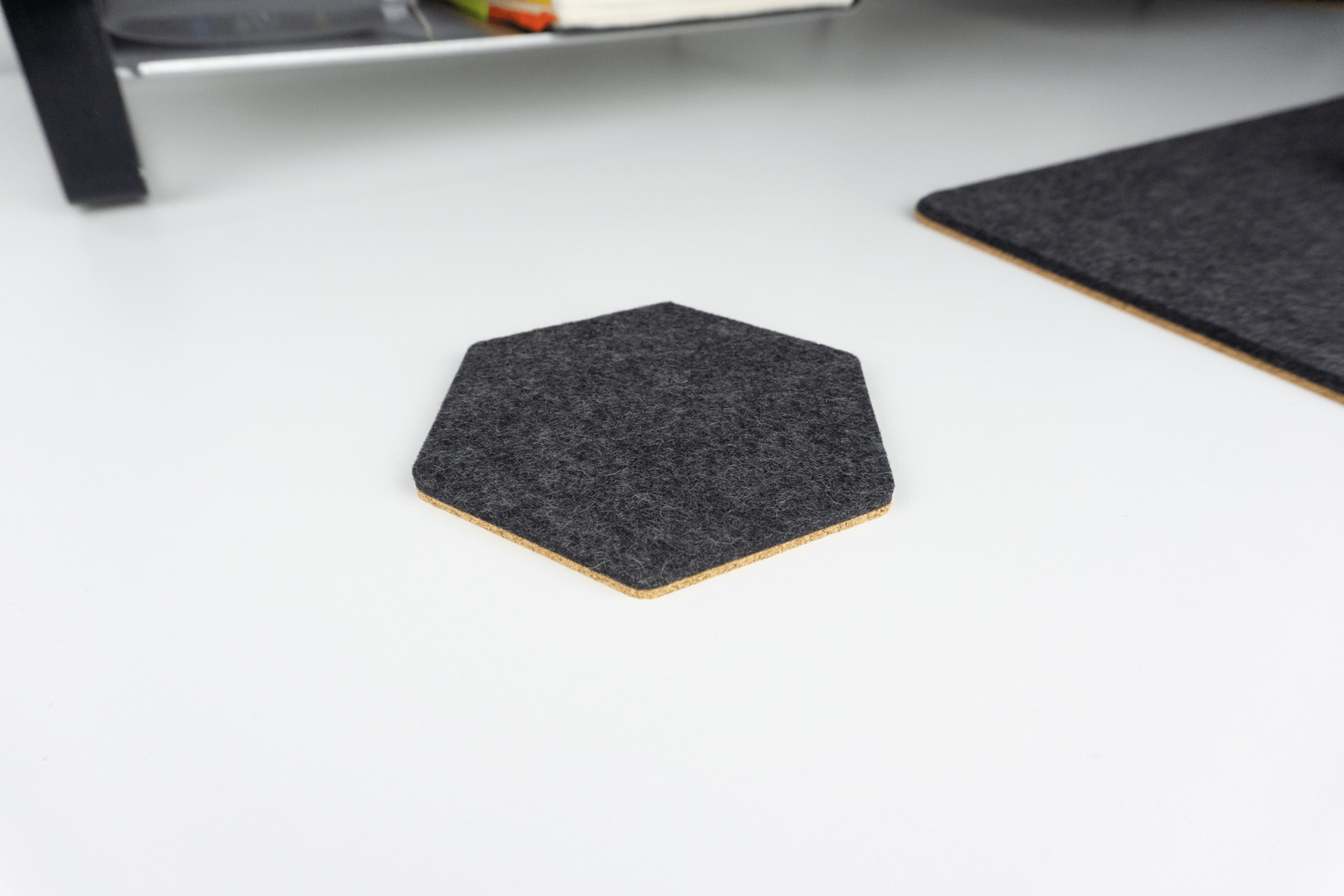 A closeup of a black hexagon coaster on a white desk next to a matching black wool and cork desk pad.