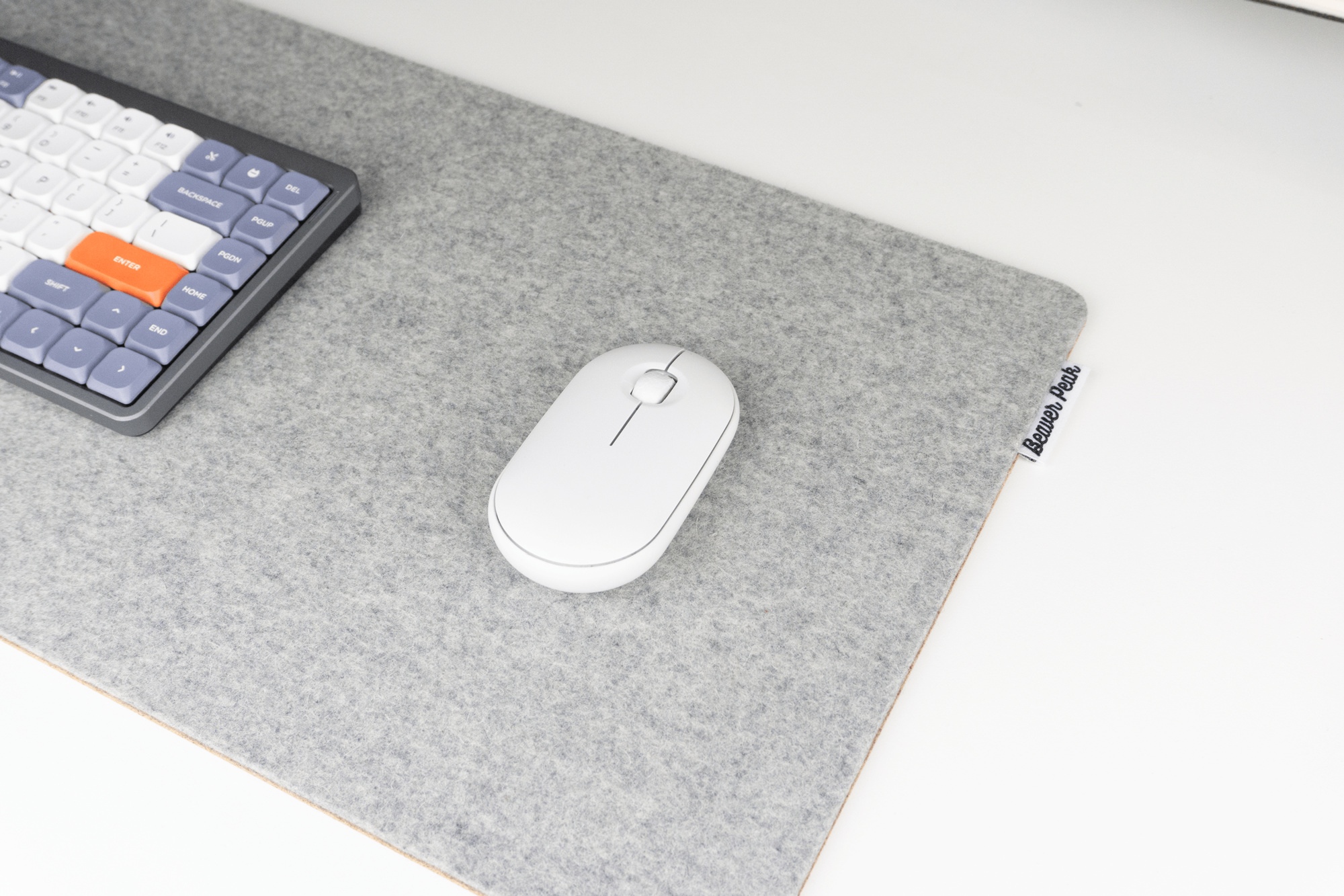 Closeup of a grey merino wool desk mat with white mouse and Nuphy air keyboard on top. The wool desk mat is on an office desk and has a BeaverPeak logo tag protruding from the right side of the mat.