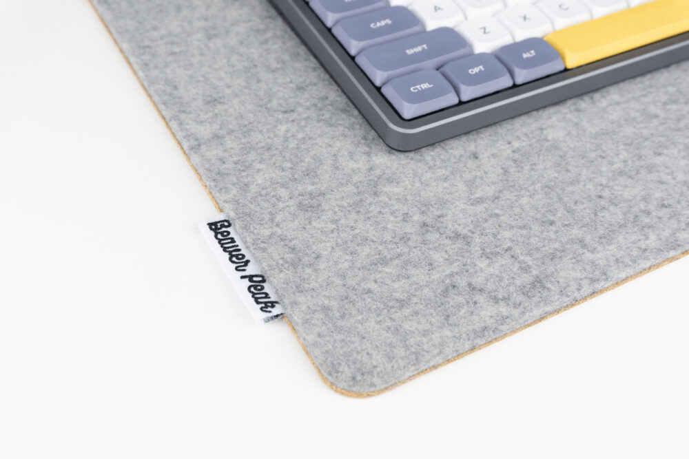 Close up of grey merino wool felt and cork desk pad with BeaverPeak logo tag on the left side and a Nuphy keyboard on top.