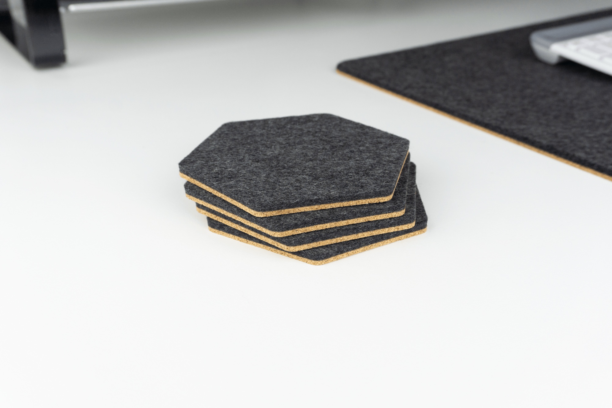 A set of 4 hexagonal desk coasters stacked on a white desk made with black merino wool and cork.