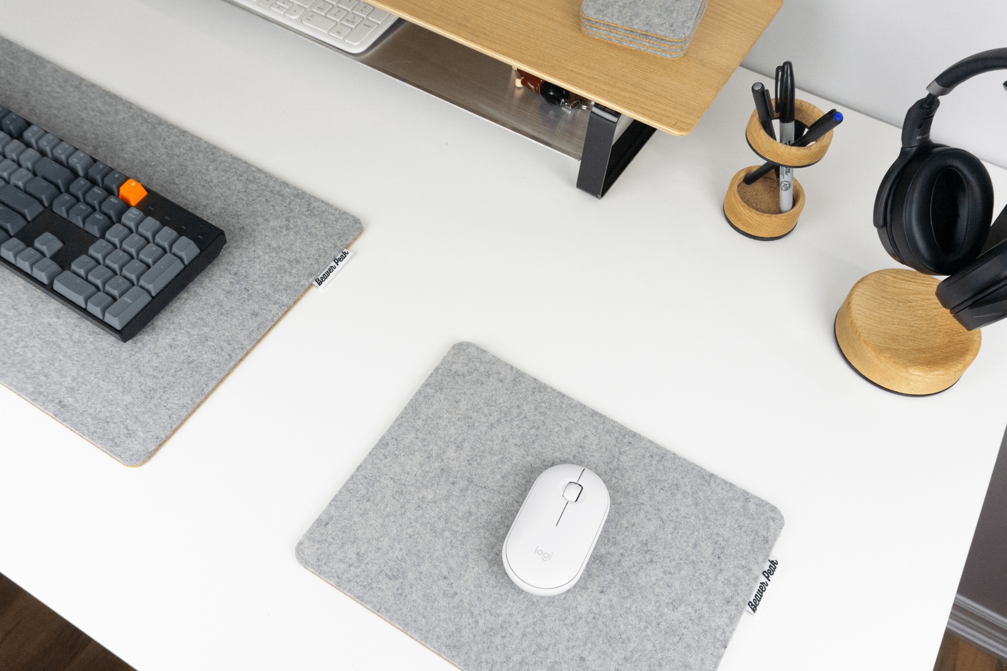 Grey wool mouse pad next to wool felt desk mat and wooden desk shelf with storage. Wooden pen holder and headphone stand at corner of desk.